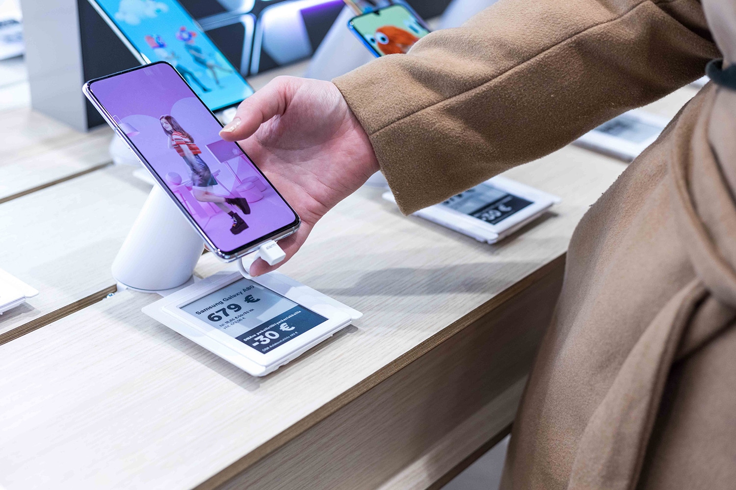 Person in a tan coat holds a smartphone at a store counter, with price cards visible nearby.