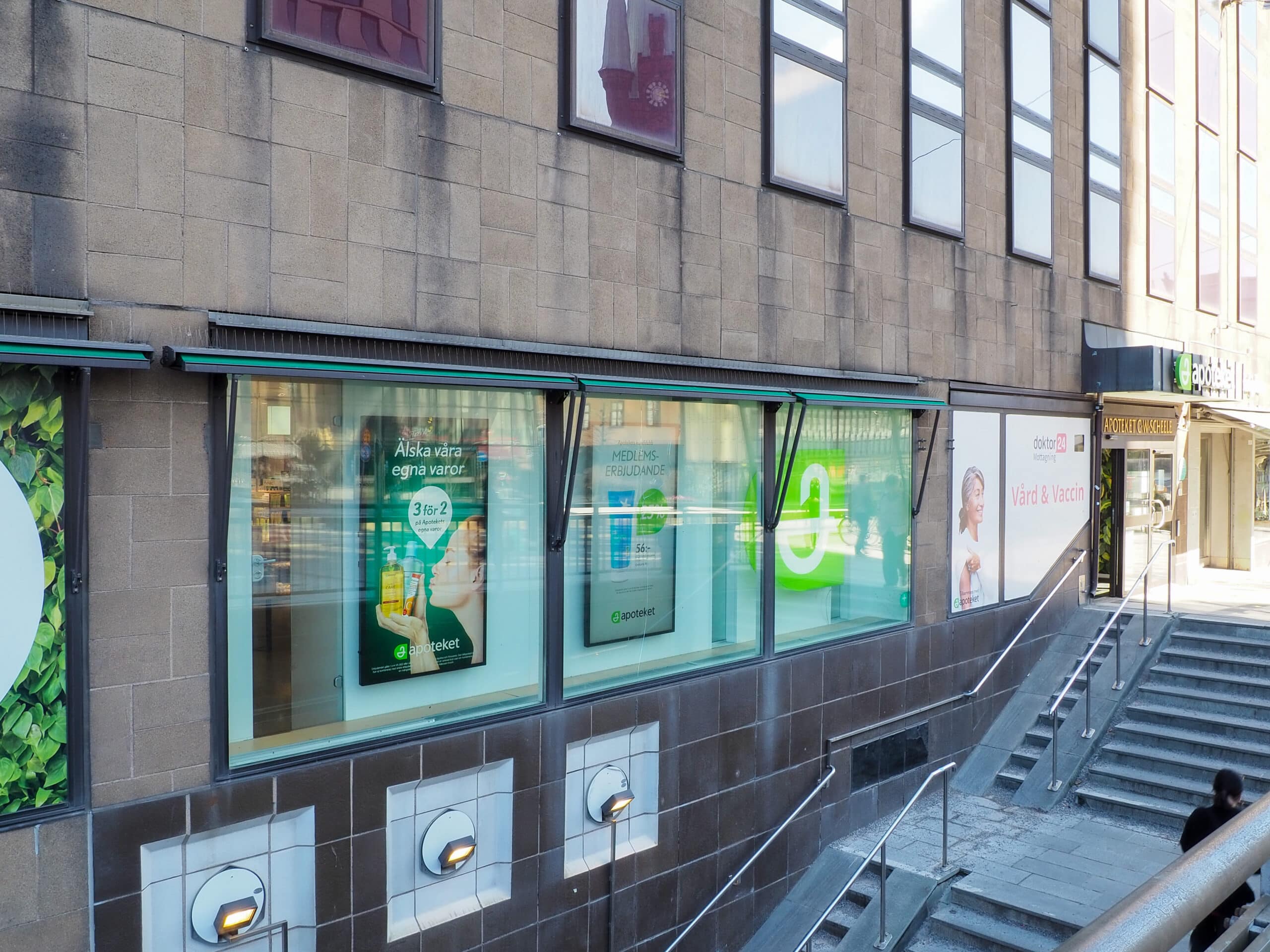 Pharmacy storefront along a sidewalk with large glass windows, green signage, and posters, set in a gray stone building with steps nearby.