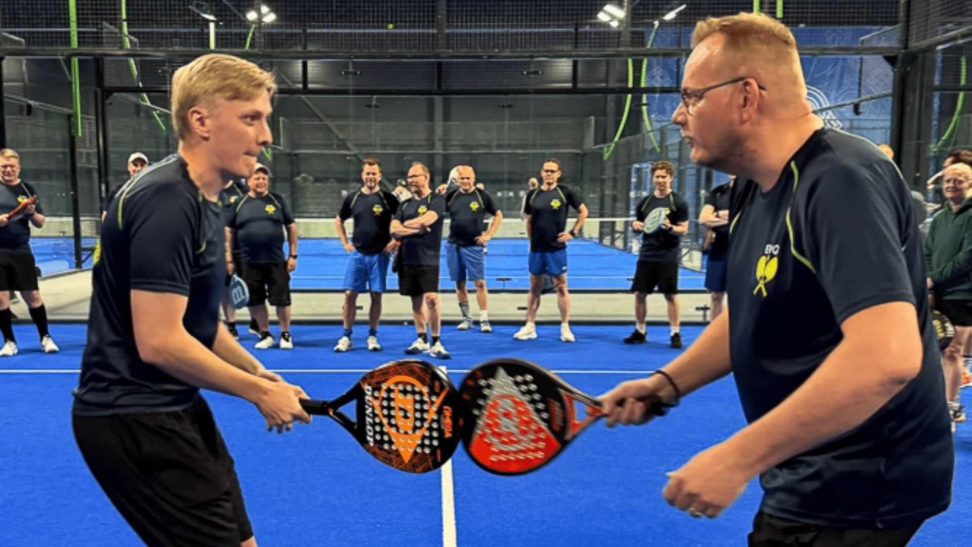 Two men on a blue indoor padel court hold their paddles together as a crowd watches in the background.